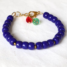 Load image into Gallery viewer, A beaded bracelet made of glossy deep blue beads, featuring a gold-tone clasp with two dangling charms, one red and one green, on a white textured fabric background.