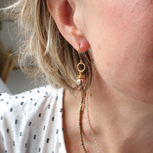 Load image into Gallery viewer, A close-up of a person wearing a delicate gold dangle earring with a small circular hoop, a pearl-like bead, and a long beaded chain, paired with a white top with black polka dots.