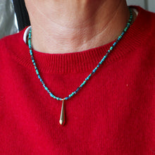 Load image into Gallery viewer, A close-up of a person wearing a beaded necklace with turquoise-blue and purple beads and a small gold teardrop pendant, paired with a red knit sweater.