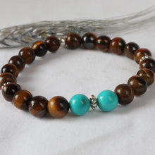 Load image into Gallery viewer, A beaded stretch bracelet featuring brown tiger's eye beads, two bright turquoise beads, and small silver spacer beads, displayed on a light gray fabric background.