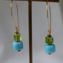 Load image into Gallery viewer, A pair of gold-tone dangle earrings with faceted light blue beads and green crystal accents, hanging from a brass display stand against a neutral gray background.