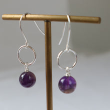 Load image into Gallery viewer, A pair of silver drop earrings with circular hoops and polished purple beads, displayed on a wooden stand against a soft gray background.