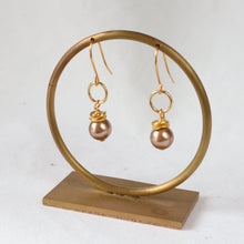 Load image into Gallery viewer, A pair of gold-tone dangle earrings with round pearl-like drops, displayed on a gold-tone circular stand with a rectangular base against a plain light background.