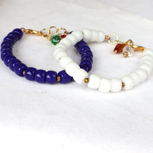 Load image into Gallery viewer, Two beaded handmade  bracelets, one in glossy deep blue and one in matte white, both with gold-tone clasp hardware and small dangling charms, displayed on a white textured background.