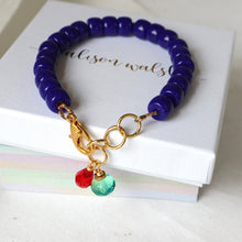 Load image into Gallery viewer, A beaded bracelet with deep blue beads, gold-tone clasp, and two dangling charms (one red, one green) resting on a white box.