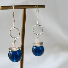 Load image into Gallery viewer, A pair of silver hoop earrings with a small pale pink bead, a silver leaf-shaped charm, and a large polished blue bead dangle, displayed on a wooden stand against a soft white background.