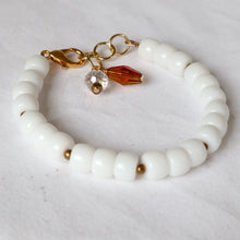 Load image into Gallery viewer, A beaded bracelet made of glossy white cylindrical beads with small gold spacer accents, a gold lobster clasp, and two charms: a faceted amber-orange crystal and a clear round crystal.