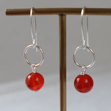 Load image into Gallery viewer, A pair of silver dangle earrings with circular wire hoops and glossy red bead drops, displayed on a wooden T-stand against a soft gray background.