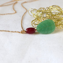 Load image into Gallery viewer, Necklace with green leaf-shaped pendant and red bead on gold chain, placed on a white fabric background.