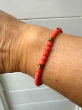 Load image into Gallery viewer, A close-up of a person's wrist wearing a beaded stretch bracelet made of coral-pink beads with small metallic gold spacer accents, set against a light gray paneled background.