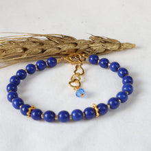 Load image into Gallery viewer, A beaded bracelet with deep blue round beads, small gold spacer details, and a gold heart-shaped clasp with a light blue faceted bead charm, displayed on a white surface with a decorative wheat stalk.