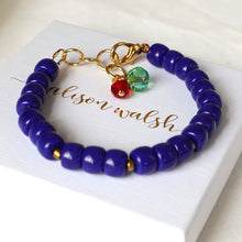 Load image into Gallery viewer, A beaded bracelet with glossy deep blue beads, gold-tone clasp hardware, and two dangling beads (one red, one green) resting on a white box with gold script.
