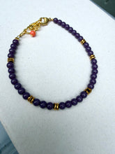 Load image into Gallery viewer, A beaded bracelet made of dark purple round beads, accented with small gold spacer beads and a gold lobster clasp, with a tiny coral-red bead charm near the clasp, displayed on a light gray textured surface.