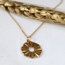 Load image into Gallery viewer, A gold-toned necklace with a textured, sunburst flower-like pendant, displayed on a white surface with a decorative gold wheat stalk in the background.