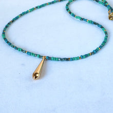 Load image into Gallery viewer, A beaded necklace featuring small turquoise and green beads with tiny gold spacer accents and a polished gold teardrop pendant, displayed on a light blue background.