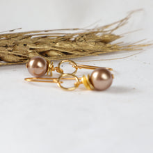 Load image into Gallery viewer, A pair of gold-toned dangle earrings with round metallic bronze beads, displayed on a white surface with dried wheat stalks in the background.