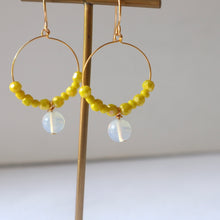 Load image into Gallery viewer, A pair of gold-plated hoop earrings with yellow beads and a shimmering opaline gemstone, displayed on a wooden stand.