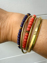 Load image into Gallery viewer, A close-up of a person's wrist wearing four stacked bracelets: two beaded bracelets in dark purple and coral red with small gold spacer beads, and two polished gold bangles, one with engraved horizontal lines and one with a textured chevron pattern.