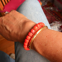 Load image into Gallery viewer, A close-up of a person's wrist wearing two coral-pink beaded bracelets with small gold accents and one wide gold bangle, set against denim jeans.