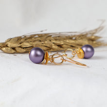 Load image into Gallery viewer, A pair of gold-tone dangle earrings with round purple pearl-like beads, displayed on a light surface with dried wheat stalks in the background.