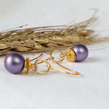 Load image into Gallery viewer, A pair of gold-tone dangle earrings with smooth, round lavender-purple pearl-like beads, displayed on a light surface with dried wheat stalks in the background.