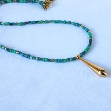 Load image into Gallery viewer, A beaded necklace featuring turquoise and teal beads with small gold spacer beads and a polished gold teardrop pendant, displayed on a light blue textured background.