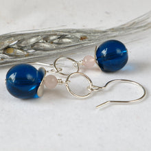 Load image into Gallery viewer, A pair of handmade dangle earrings featuring glossy deep blue glass beads, small pale pink beads, and silver wire hooks, displayed on a textured white surface with a silver decorative prop.
