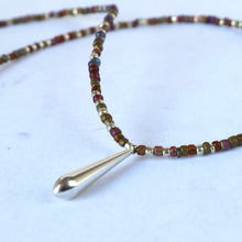 Load image into Gallery viewer, A beaded necklace with small metallic and earthy beads and a polished silver teardrop pendant, photographed on a light blue background.