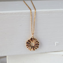 Load image into Gallery viewer, A gold chain necklace with a textured, sunburst-style oval pendant, resting on a white painted wooden surface.
