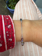 Load image into Gallery viewer, A close-up of a person's wrist wearing a delicate silver-toned bangle bracelet with small faceted beads in purple, green, and white. The person is wearing a red patterned sleeve and light pink striped pants, with a potted plant visible in the background.