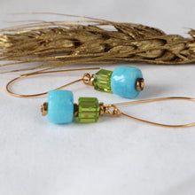 Load image into Gallery viewer, Gold-tone wire drop earrings with a turquoise-blue round bead, a faceted lime-green cube bead, and a small gold spacer, displayed on a white surface with a decorative gold wheat stem in the background.