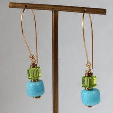 Load image into Gallery viewer, A pair of gold-tone dangle earrings with light blue marbled beads, faceted green crystal accents, and long curved gold hooks, displayed on a brass T-stand against a neutral gray background.